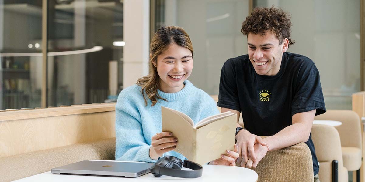 Two students look at an open notebook on a balcony in a modern building on campus during the day. One stands and the other sits down.