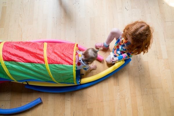 A baby crawls through a play tunnel while an older child watches and helps.