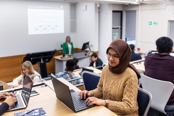 A student using a laptop during a lecture. There are other students on laptops in view and a lecturer is presenting in the background.