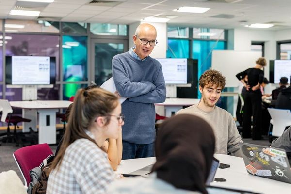 A group of students in a classroom sat at a desk with a tutor stood up talking to them.