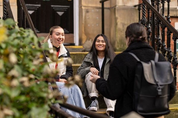 Three students chatting and smiling. Two are sat on steps outside an old characterful university building. The other is stood with them.