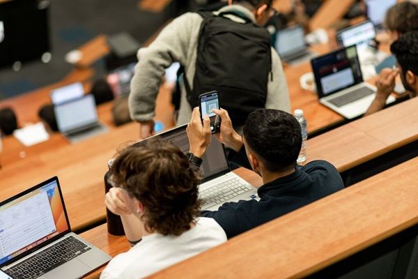 Several students seated with their laptops or taking seats in a lecture theatre, viewed from behind.